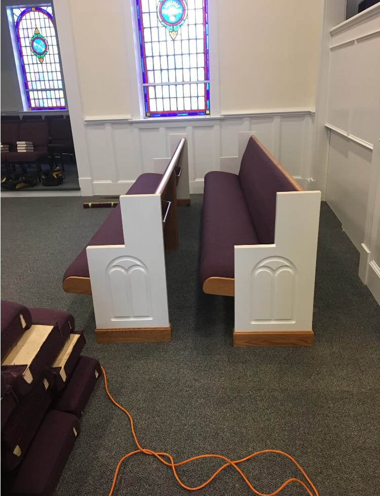 New Pews Installation – Initial Placement (2018) New pew installation underway at Rawls Baptist Church in 2018, featuring white and wood-trimmed ends with plum upholstery beside stained glass windows.