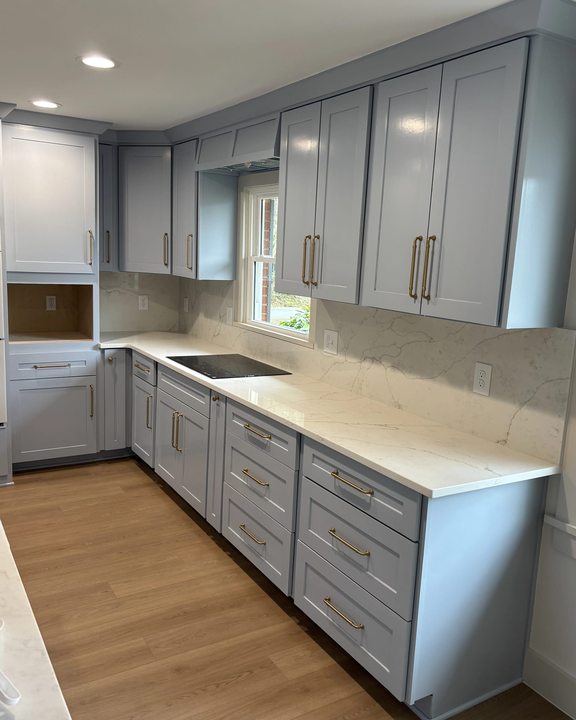 Sleek modern kitchen with light blue shaker cabinets, brass hardware, white quartz countertops, and integrated cooktop beneath a large window.